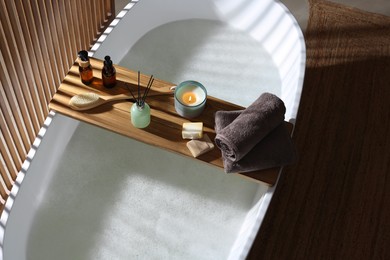 Photo of Wooden bath tray with candle, air freshener and bathroom amenities on tub indoors, above view