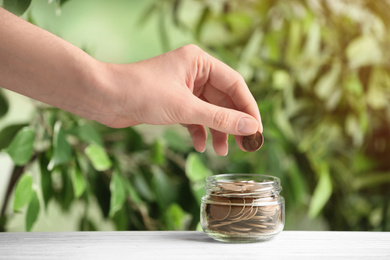 Photo of Woman putting coin into jar on white wooden table against blurred green background, closeup. Money savings