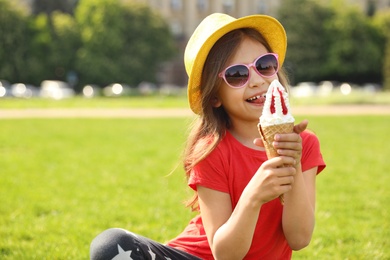 Cute little girl with delicious ice cream sitting on green grass outdoors, space for text