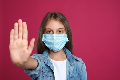 Photo of Little girl in protective mask showing stop gesture on crimson background, space for text. Prevent spreading of coronavirus