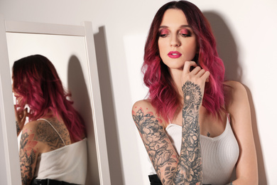Photo of Beautiful woman with tattoos on body near mirror indoors