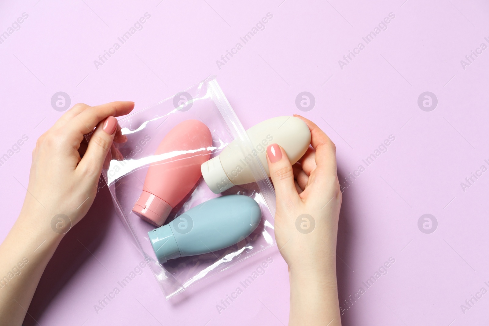 Photo of Cosmetic travel kit. Woman putting small bottle with personal care product into plastic bag against violet background, top view