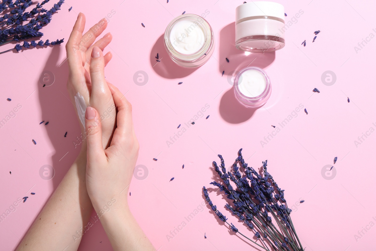 Photo of Woman applying hand cream and lavender flowers on pink background, top view