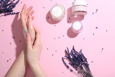 Woman applying hand cream and lavender flowers on pink background, top view