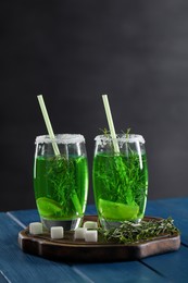 Photo of Glasses of homemade refreshing tarragon drink, sprigs and sugar cubes on blue wooden table, space for text