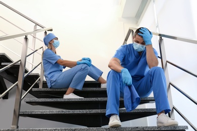 Photo of Exhausted doctors sitting on stairs indoors. Stress of health care workers during COVID-19 pandemic