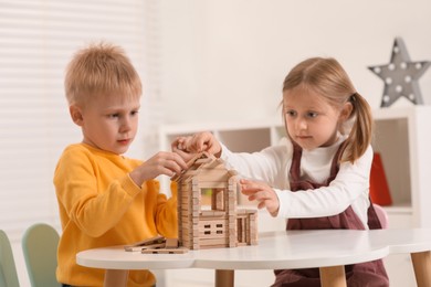 Little boy and girl playing with wooden house at white table indoors. Children's toys