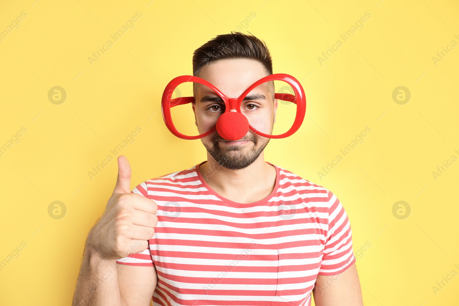 Photo of Young man with party glasses and clown nose on yellow background. April fool's day