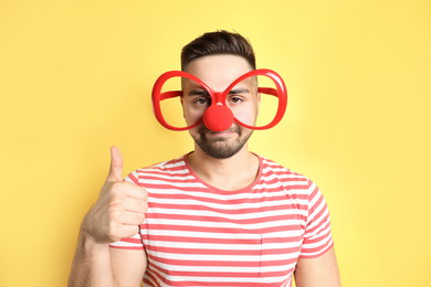 Photo of Young man with party glasses and clown nose on yellow background. April fool's day