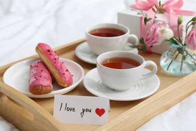 Photo of Tasty breakfast served in bed. Delicious eclairs, tea, gift box, flowers and card with phrase I Love You on tray