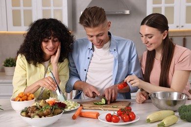 Photo of Friends cooking healthy vegetarian meal at white marble table in kitchen