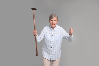 Angry senior woman with walking cane on gray background