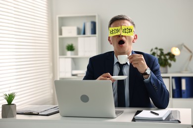 Man with fake eyes painted on sticky notes holding cup of drink and yawning at workplace in office