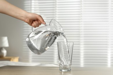 Photo of Woman pouring water from jug into glass at wooden table indoors, closeup
