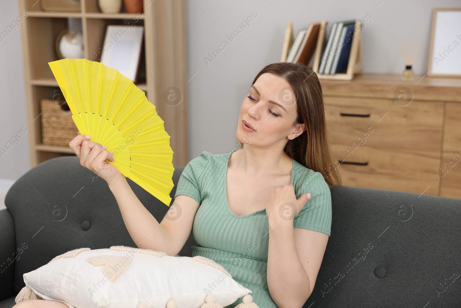 Photo of Woman waving yellow hand fan to cool herself on sofa at home