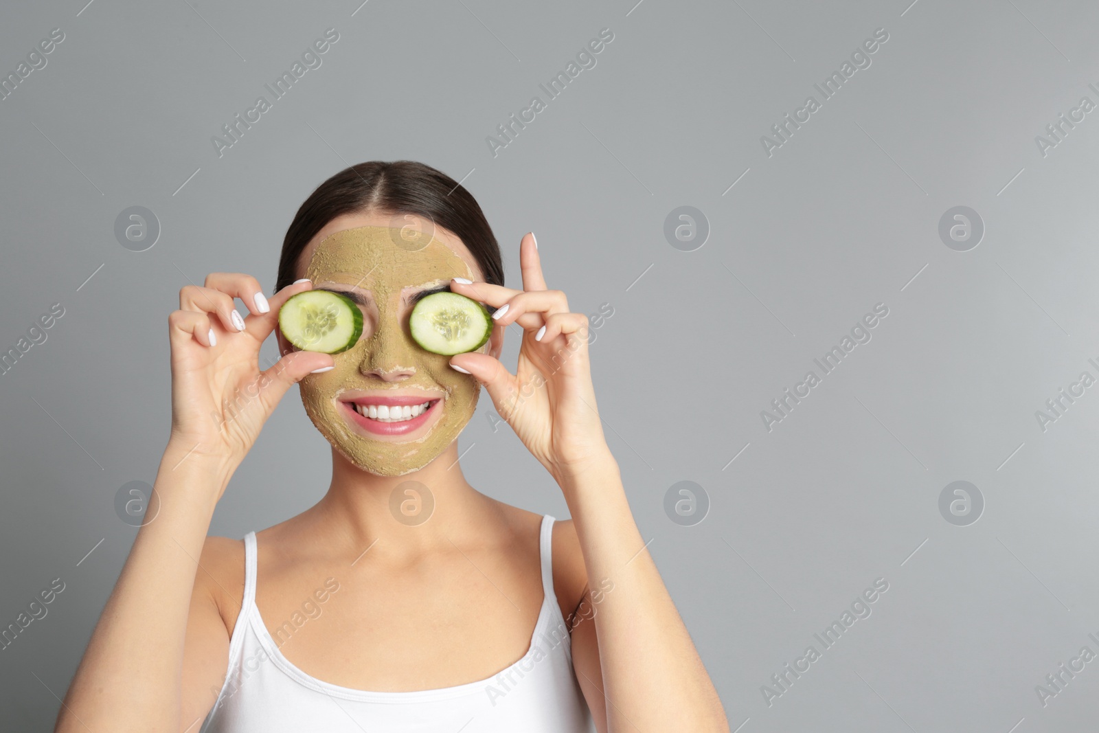 Photo of Young woman with clay mask on her face holding cucumber slices against grey background. Space for text