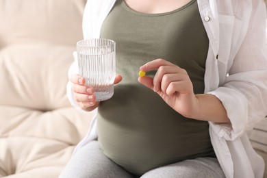 Photo of Pregnant woman holding pill and glass with water on sofa, closeup