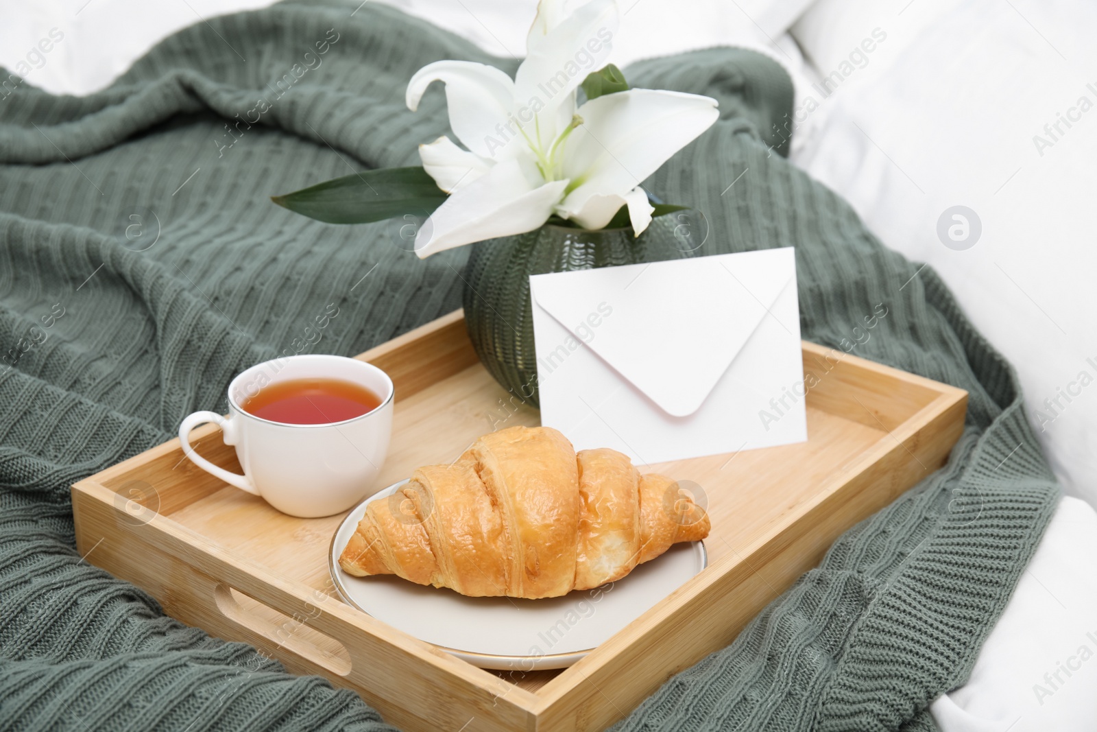 Photo of Tray with tasty croissant, cup of tea and flower on bed