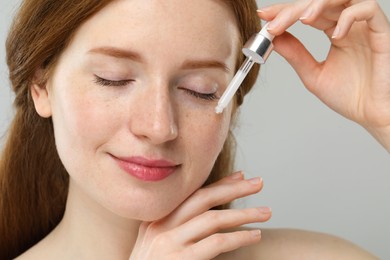 Beautiful woman with freckles applying cosmetic serum onto her face against grey background, closeup