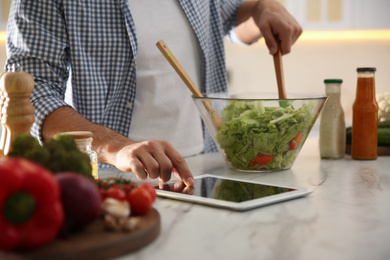Photo of Man with tablet cooking salad at table in kitchen, closeup