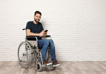 Photo of Young man with smartphone in wheelchair near brick wall indoors. Space for text
