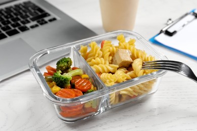 Photo of Container with tasty food, fork and laptop on white wooden table, closeup. Business lunch
