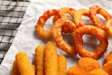 Tasty chicken nuggets, fried onion rings, cheese sticks and ketchup on parchment paper, closeup