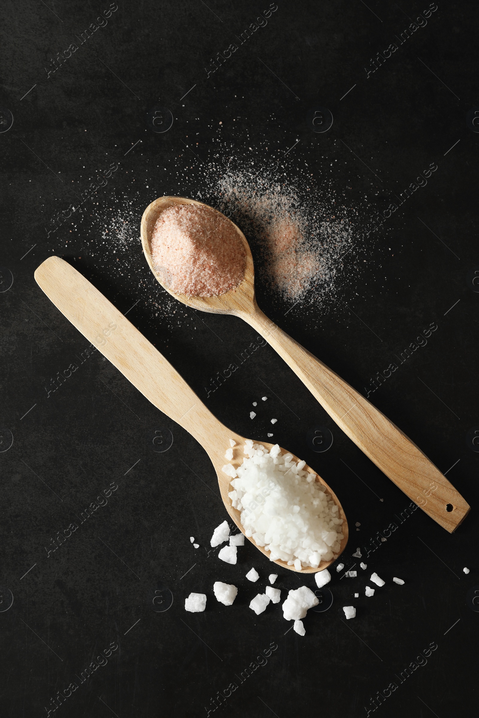 Photo of Different types of organic salt in spoons on black table, flat lay