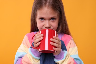 Photo of Cute girl drinking beverage from red ceramic mug on orange background