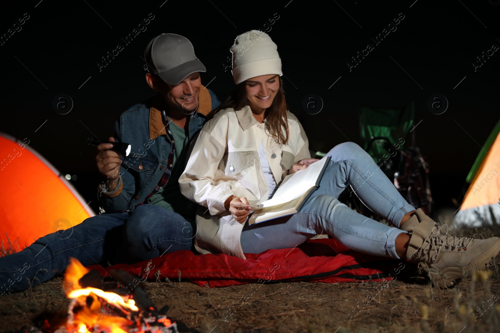 Photo of Couple with flashlight reading book near bonfire at night. Camping season