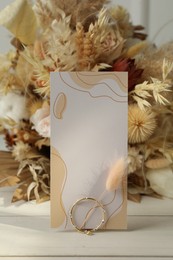 Blank invitation card, ring, dry leaves and flowers on white wooden table