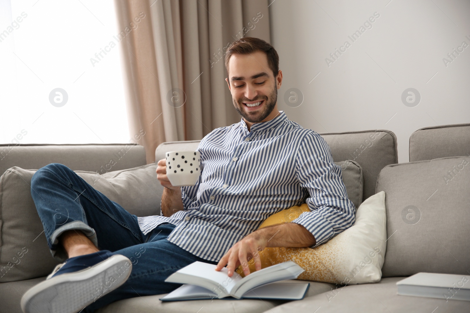 Photo of Handsome young man with cup of coffee reading book at home