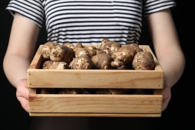 Woman holding wooden crate with Jerusalem artichokes on black background, closeup