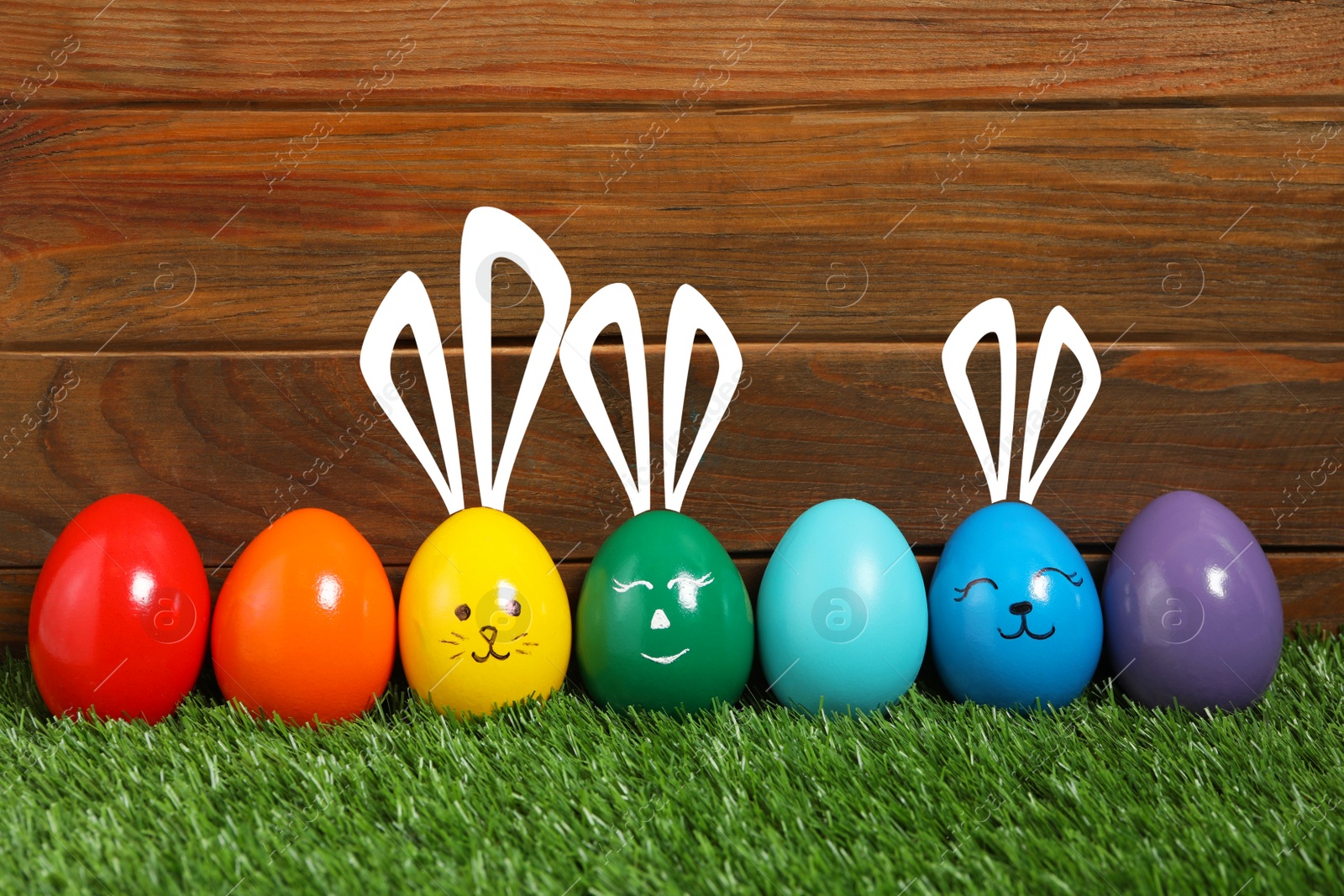 Image of Several eggs with drawn faces and ears as Easter bunnies among others on green grass against wooden background