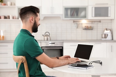 Handsome young man working with laptop at table in kitchen