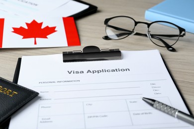 Photo of Visa application form for immigration, passport, Canadian flag and glasses on wooden table, closeup