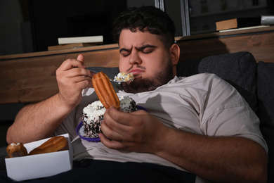 Photo of Depressed overweight man eating sweets in living room at night