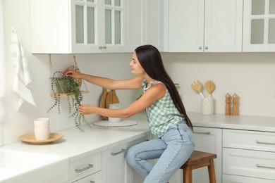 Photo of Beautiful young woman taking care of houseplant while sitting on stool in kitchen