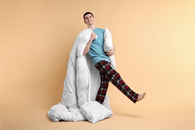 Happy man in pyjama holding pillow and blanket on beige background