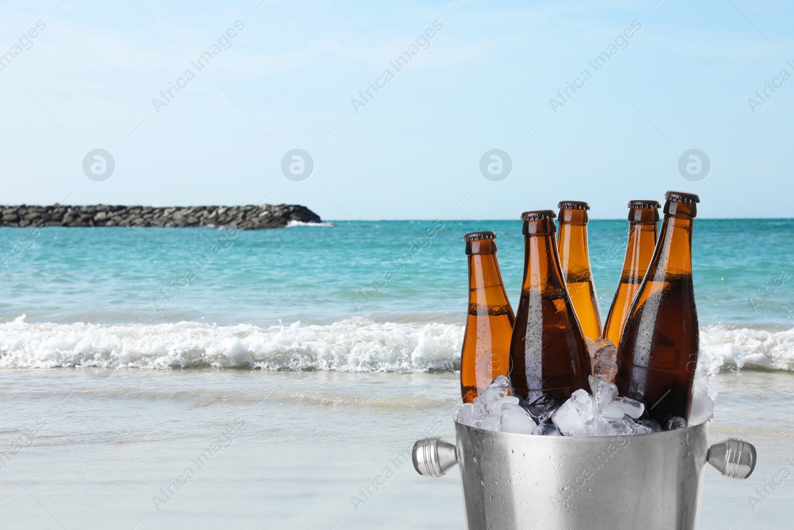 Image of Bottles of beer with ice cubes in metal bucket against ocean and sandy beach. Space for text