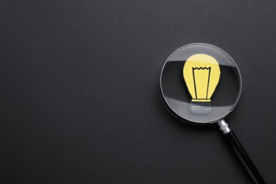 Photo of Magnifying glass over paper light bulb on dark background, top view. Space for text