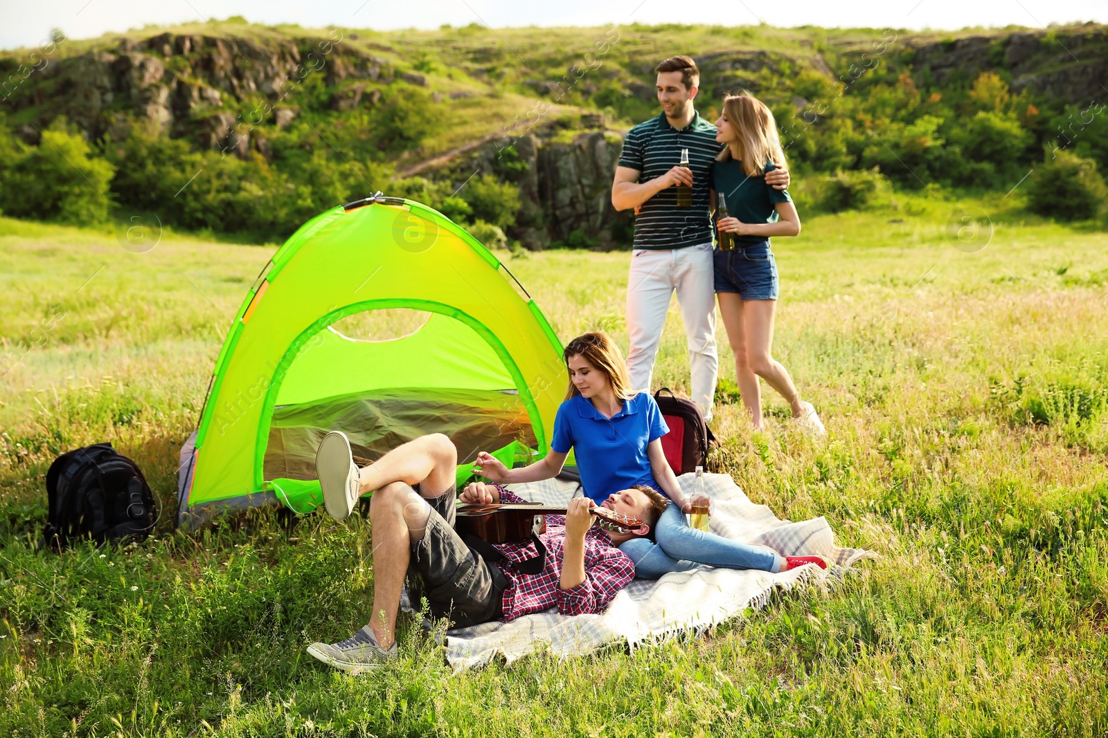 Photo of Group of young people resting with beer and guitar near camping tent in wilderness