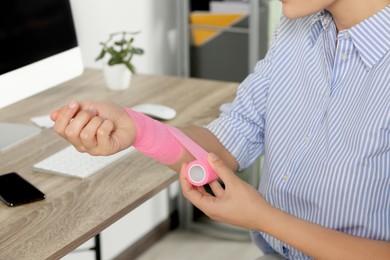 Photo of Young woman applying medical bandage onto wrist in office, closeup