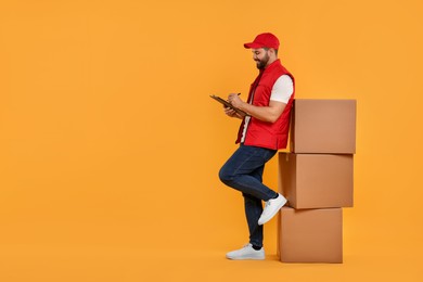 Photo of Happy young courier with clipboard and stack of parcels on orange background. Space for text