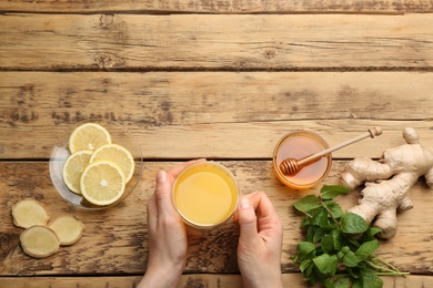 Woman holding cup of immunity boosting drink at wooden table with ingredients, top view. Space for  text