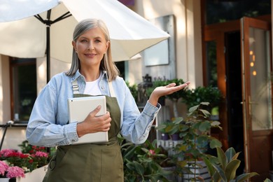 Photo of Happy business owner with tablet inviting to come into her flower shop outdoors, space for text