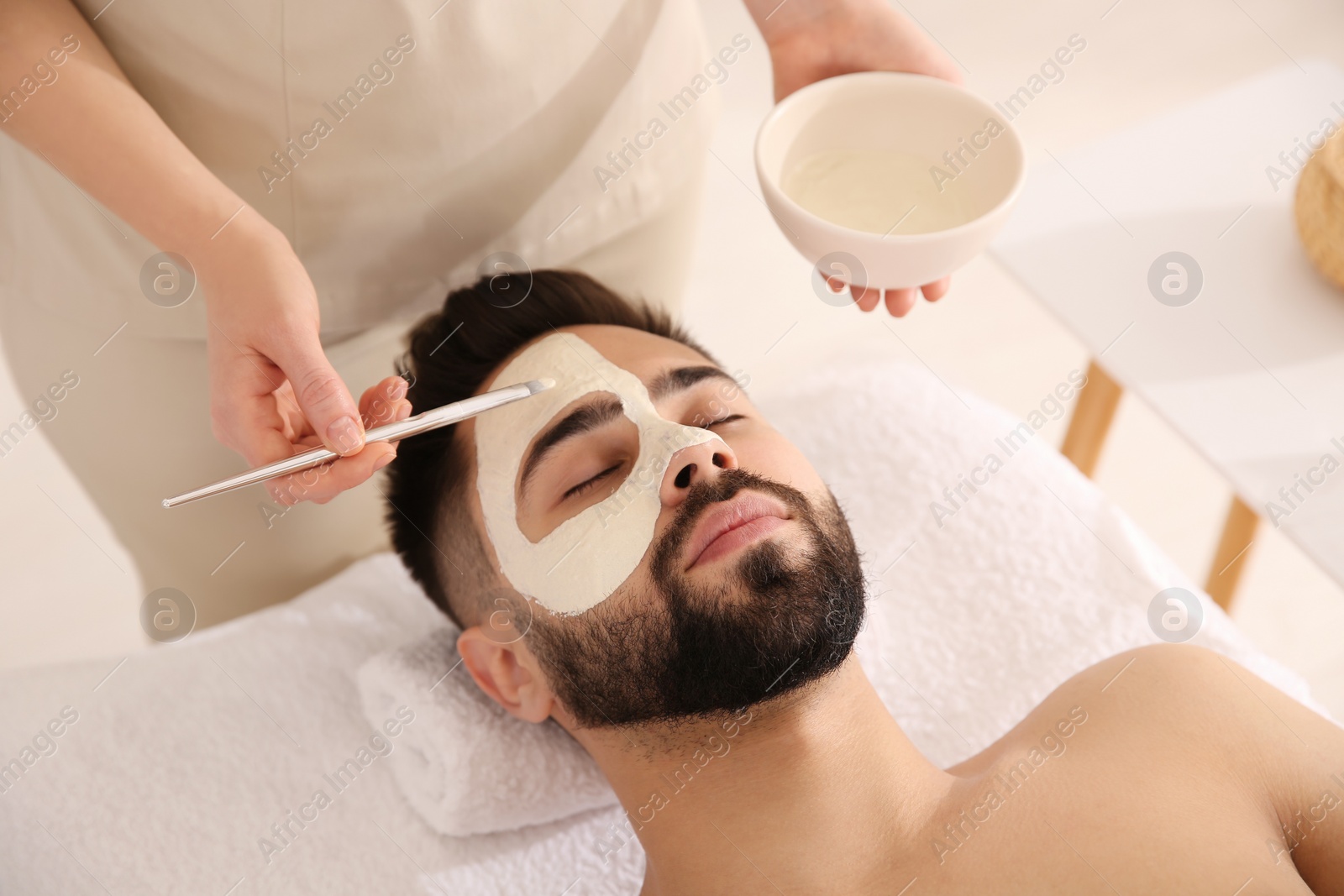 Photo of Cosmetologist applying mask on man's face in spa salon