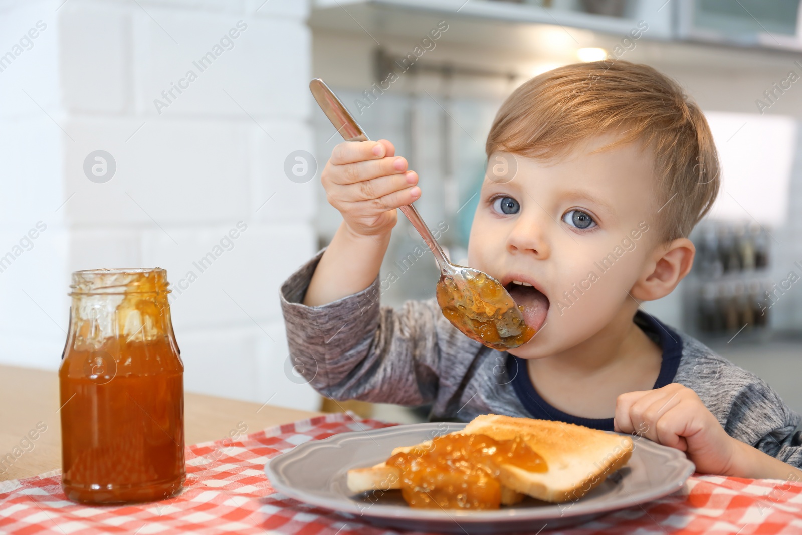 Photo of Little boy eating jam at table in kitchen