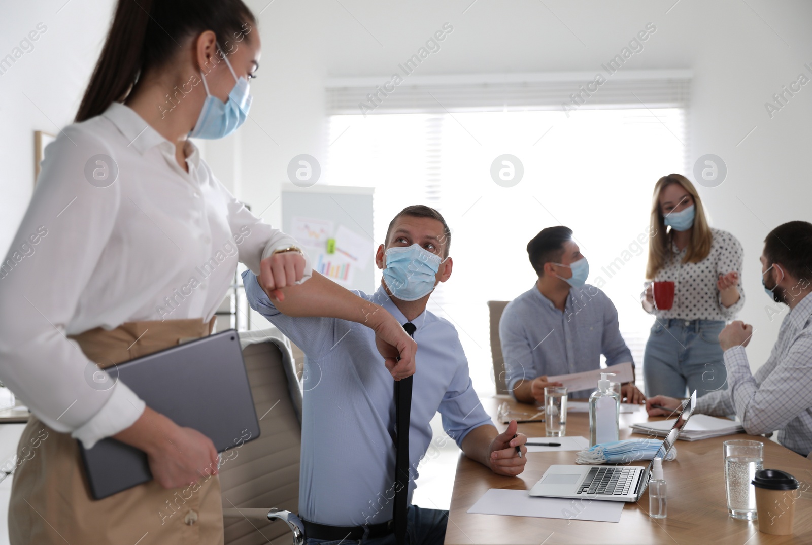 Photo of Coworkers with protective masks making elbow bump in office. Informal greeting during COVID-19 pandemic