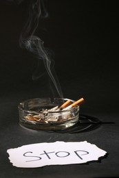 Photo of Ashtray with burnt cigarettes and word Stop written on paper on black table. No smoking concept